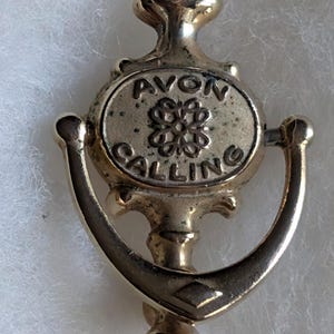 May include: A gold-toned metal brooch in the shape of a door knocker. The central oval plaque displays the text AVON CALLING with a decorative knot motif in the centre. The item has a polished finish and includes a hinged, U-shaped swinging component.