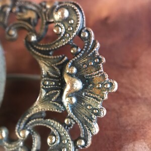 Puede incluir: Un broche de metal dorado con un dise&ntilde;o intrincado y ornamentado. El broche presenta un elemento central en forma de abanico con una superficie texturizada en relieve.