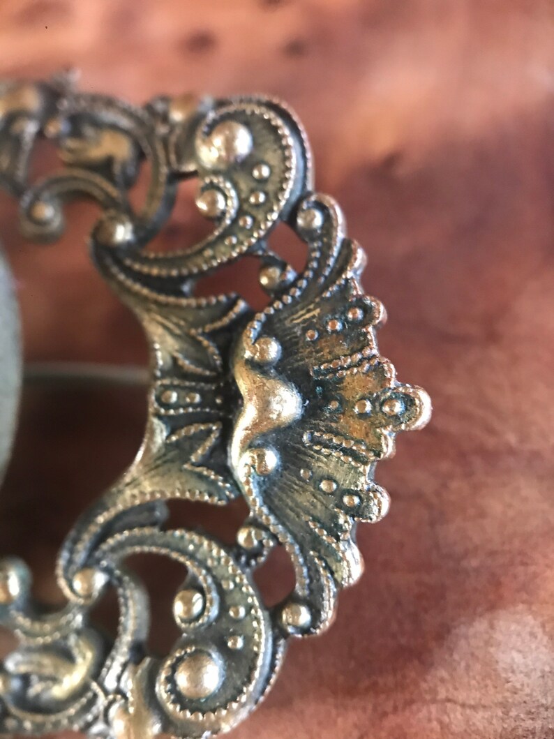 Puede incluir: Un broche de metal dorado con un dise&ntilde;o intrincado y ornamentado. El broche presenta un elemento central en forma de abanico con una superficie texturizada en relieve.