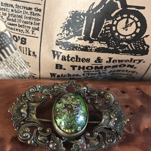 Puede incluir: Un broche vintage con un marco de plata adornado y una piedra ovalada de cabuj&oacute;n verde. La piedra tiene un patr&oacute;n de m&aacute;rmol con motas doradas.