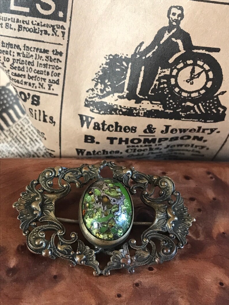 Puede incluir: Un broche vintage con un marco de plata adornado y una piedra ovalada de cabuj&oacute;n verde. La piedra tiene un patr&oacute;n de m&aacute;rmol con motas doradas.