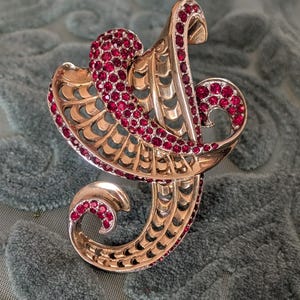 May include: A gold-toned brooch with a swirling, abstract design. The brooch is embellished with numerous small, round, red gemstones. The design features openwork and curved elements, creating an elegant and ornate appearance. The brooch is likely vintage or antique.