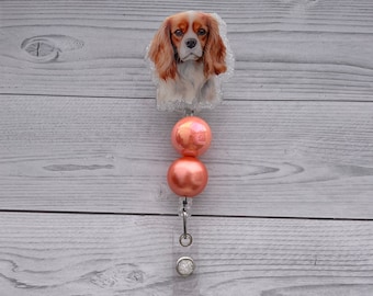 May include: A badge reel featuring a Cavalier King Charles Spaniel illustration on a glittery acrylic topper, accented with two round, glossy peach-colored beads, a metal swivel clip, and a clear plastic strap with a silver-toned snap button closure.