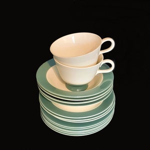 May include: A set of teacups and saucers in teal and white. Two white teacups are stacked on top of a pile of teal and white saucers. The teacups have a rounded shape and a handle. The saucers have a teal rim and a white centre.