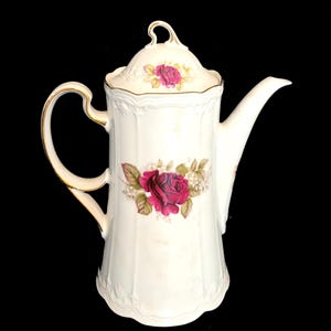 May include: A white porcelain coffee pot with a lid, decorated with a large red rose floral design. The pot has a gold handle and trim, and a curved spout. The design includes a smaller rose on the lid.