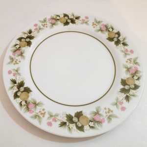 May include: A white ceramic dinner plate with a green and brown floral pattern featuring pink flowers, green leaves, and yellow fruit.