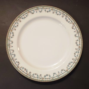 May include: A white ceramic plate with a green and gold floral border. The plate has a simple design with a white center and a decorative rim.