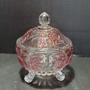 Op de afbeelding: Een glazen snoepvaas met deksel, met een druivenrankenontwerp met rode en gouden accenten. De vaas heeft drie poten en is perfect om snoep, noten of andere lekkernijen te serveren.