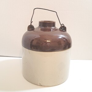 May include: A brown and white glazed stoneware crock with a wire handle. The crock has a wide mouth and a rounded body.