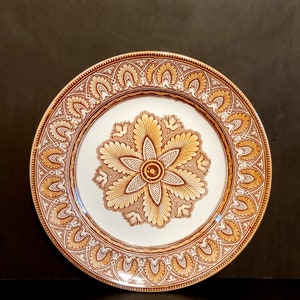 May include: A white ceramic plate with a brown and gold floral design. The plate has a raised rim and a scalloped edge.