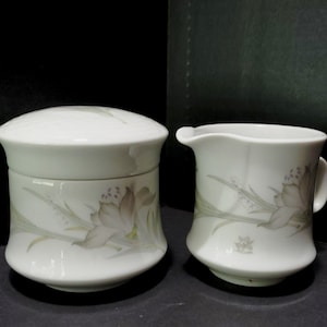 May include: A white ceramic sugar bowl with a lid and a creamer. Both pieces are decorated with a floral pattern in shades of gray and green. The sugar bowl is approximately 4 inches tall, and the creamer has a handle.