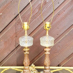 May include: A pair of gold table lamps with clear glass shades and ornate bases. The shades have a textured, diamond-like pattern. The bases are decorated with a leaf design.