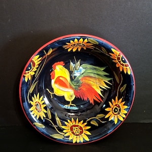 May include: A black ceramic bowl with a red rim features a hand-painted design of a rooster and sunflowers. The rooster is yellow, red, and green, and the sunflowers are yellow and brown.