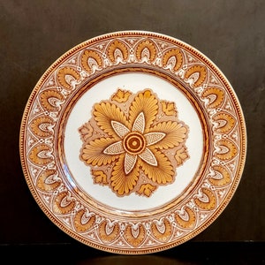May include: A white ceramic plate with a brown and gold floral design. The plate has a decorative border with a repeating pattern of stylized leaves and flowers.
