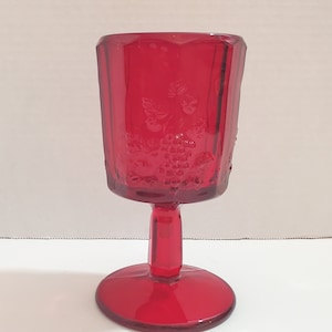 May include: A red glass goblet with a grape design. The goblet has a stemmed base and a wide, round bowl.