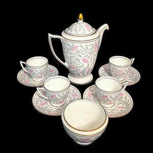 May include: A porcelain tea set featuring a tall teapot with a gold-toned finial, four teacups with matching saucers, and a sugar bowl. Each piece is decorated with a repeating pattern of pink flowers and grey foliage on a white background.