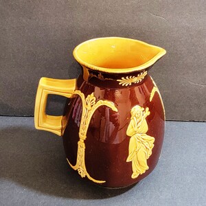 May include: A brown ceramic pitcher with a yellow glaze and a raised design of a woman in a flowing gown. The pitcher has a handle and a wide mouth.