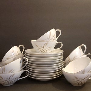 May include: A set of white teacups and saucers with a delicate wheat design in gold. The cups and saucers are stacked neatly, showcasing the elegant pattern.