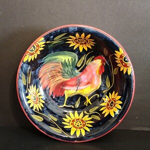 May include: A black ceramic bowl with a red, yellow and green rooster design. The bowl is decorated with sunflowers and has a red rim.
