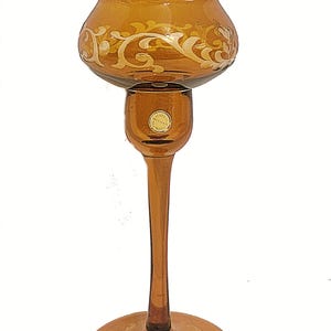 May include: Amber-colored glass candle holder with a round base and a tall, slender stem. The bowl-shaped top and base feature a white, ornate floral design. The candle holder is approximately 12 inches tall.