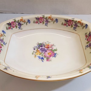May include: A white ceramic serving bowl with a gold rim and floral pattern. The bowl has a square shape with rounded corners and features a bouquet of pink, purple, and yellow flowers in the center.