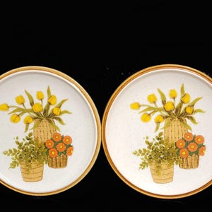 May include: Two ceramic plates with a floral design featuring yellow tulips and orange flowers in baskets. The plates have a beige background with a speckled pattern.
