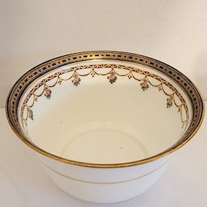 May include: White porcelain bowl with a blue and gold rim. The rim is decorated with a repeating pattern of gold dots and a gold garland with pink roses.