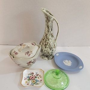 Könnte beinhalten: Ein Set aus vier Vintage-Keramik- und Glasartikeln. Ein grün-brauner, gesprenkelter Krug, eine weiße Schüssel mit Deckel und Blumendesigns, ein blauer und weißer Dekorationsteller und ein grüner Glasdeckel.