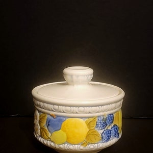 May include: A white ceramic sugar bowl with a lid, decorated with a colorful fruit design. The design features yellow lemons, blue plums, green leaves, and purple berries.