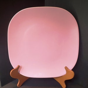 May include: A pink ceramic dinner plate on a wooden stand. The plate is round with slightly rounded edges.