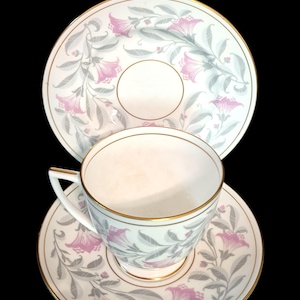 May include: A white porcelain teacup and saucer set featuring a delicate floral pattern with pink trumpet-shaped flowers and grey leaves. Both the cup and saucer are accented with a thin gold rim, and the cup includes a small, angled handle.