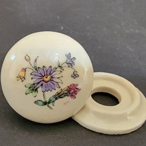 May include: A white ceramic knob with a floral design featuring purple, yellow, and pink flowers. The knob has a ring base.