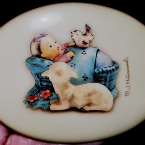 May include: A white oval ceramic plate with a painted scene of a child in a blue dress holding a white lamb. A small bird is perched on the child's shoulder. The plate is signed "M.J. Hummel" in the lower right corner.