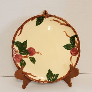 May include: A vintage ceramic plate with a brown twig design and pink apples with green leaves. The plate is on a wooden stand.