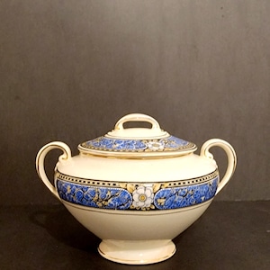 May include: A white ceramic sugar bowl with a lid, featuring a blue and gold floral pattern. The bowl has two handles and a round base.