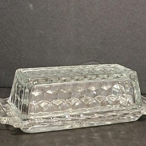 May include: A clear glass butter dish with a diamond pattern. The dish has a lid and a base with a handle on each side.