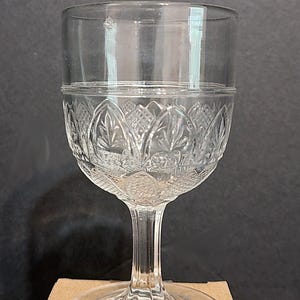 May include: A clear glass goblet with a patterned bowl and a slender stem. The bowl has an embossed design of leaves and floral elements. The goblet sits on a round base. Suitable for serving drinks, it is made of clear glass.