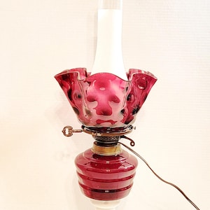 May include: A vintage oil lamp with a white base and a burgundy glass shade with a ruffled edge. The shade has a textured, bubbled design.