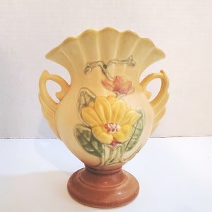Könnte beinhalten: Eine gelbe und braune Keramikvase mit einem gewellten Rand und zwei Griffen. Die Vase ist mit einem Blumendesign verziert, das gelbe, rosa und grüne Blumen zeigt.
