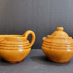 May include: A brown ceramic creamer and sugar bowl set with a rustic design. The creamer has a handle and a spout, while the sugar bowl has a lid.