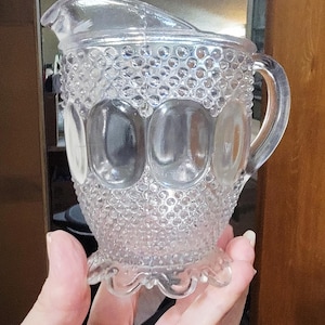 May include: A clear glass pitcher with a textured surface and a scalloped base. The pitcher has a handle and a wide mouth.
