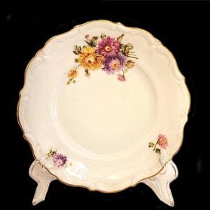 May include: A white porcelain plate with a gold rim and a floral design featuring yellow, pink, and purple flowers. The plate has a scalloped edge.
