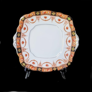 May include: A square, white ceramic cake plate with a raised, scalloped edge. The plate has a colorful floral pattern in shades of orange, red, green, and black.