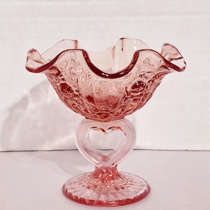 May include: A pink glass pedestal bowl with a ruffled rim and a heart-shaped stem. The bowl has a textured floral pattern and sits on a round base. The bowl is approximately 15 cm tall.