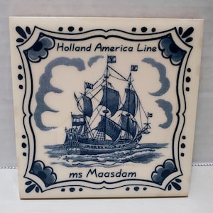 May include: A white ceramic tile with a blue and white design featuring a ship with the text "Holland America Line" and "ms Maasdam".