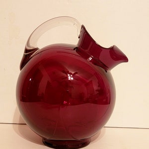 Könnte beinhalten: Eine tiefrote Glaskanne mit einem durchsichtigen Glasgriff. Die Kanne hat einen runden Körper und einen breiten Ausguss.