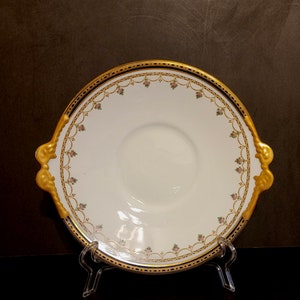 Peut inclure: Un plat de service en porcelaine blanche avec des bordures dorées et bleues. Le plat a un bord festonné et deux poignées dorées. Le plat est décoré d'un motif floral.