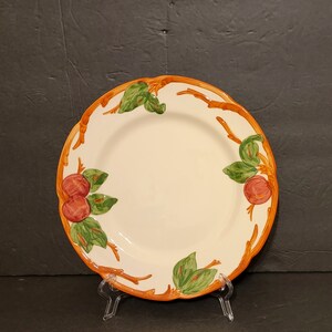 May include: A white ceramic dinner plate with a brown, orange, and green apple design. The plate has a scalloped edge and is decorated with stylized branches.