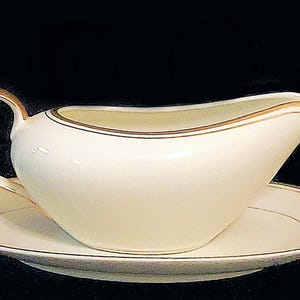 Könnte beinhalten: Cremefarbene Sauciere mit passender Untertasse. Die Porzellanstücke haben einen goldenen Rand an den Kanten und am Griff. Die Sauciere hat einen gebogenen Ausguss und einen abgerundeten Griff. Schwarzer Hintergrund.