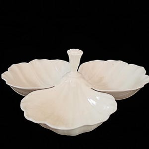 May include: A white, three-section serving dish with a central handle. The dish has a petal-like design, with each section resembling a stylized flower. The serving dish is made of ceramic and is set against a black background.